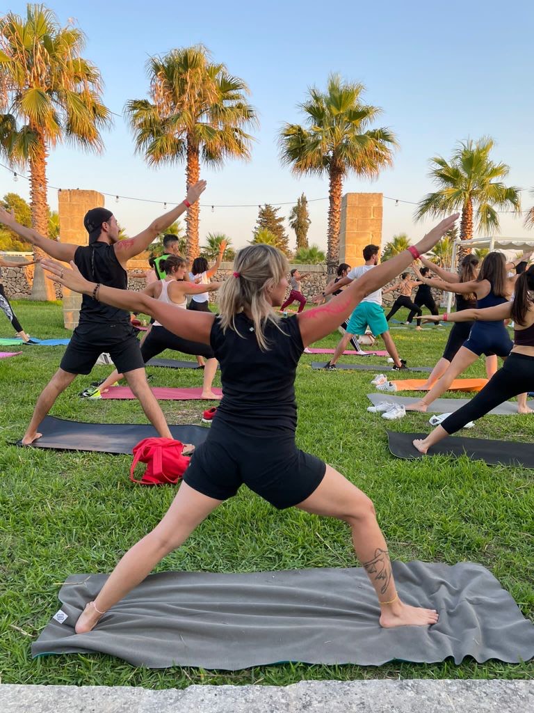 Nicoletta in posizione del guerriero durante una lezione di yoga all'aperto, su un prato tra le palme, con il gruppo che pratica dietro di lei